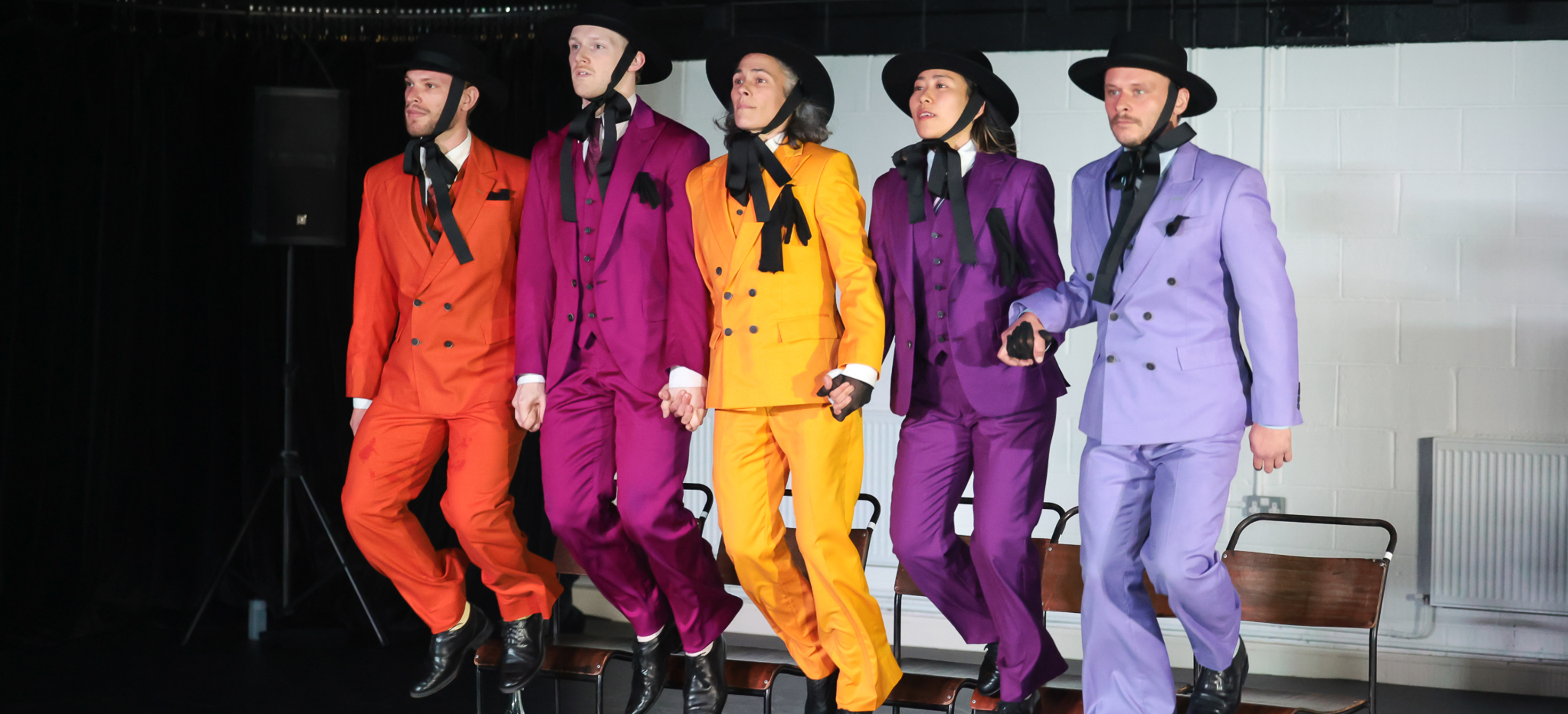 Left to right: five performers wearing orange, pink, yellow, purple and lilac suits jumping off chairs on a stage